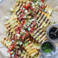 A platter of yellow squash, halved and grilled topped with bacon pieces, blue cheese, and balsamic.