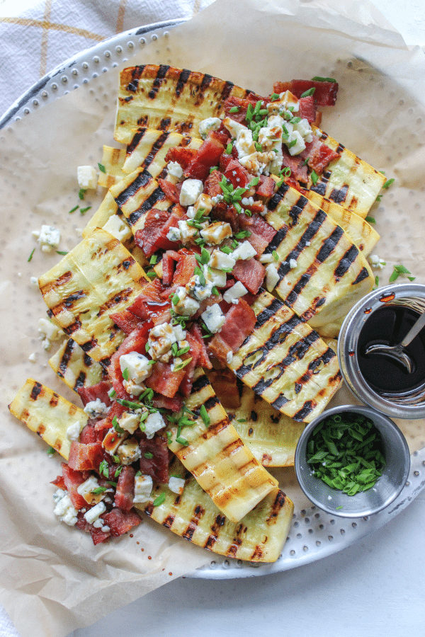 A platter of yellow squash, halved and grilled topped with bacon pieces, blue cheese, and balsamic.