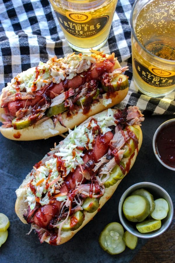 Two pulled pork hot dogs on a table with a beer.