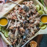 A platter of pulled pork, pickles, mustard, and forks on a table.
