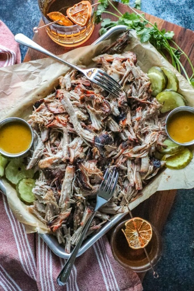 A platter of pulled pork, pickles, mustard, and forks on a table.