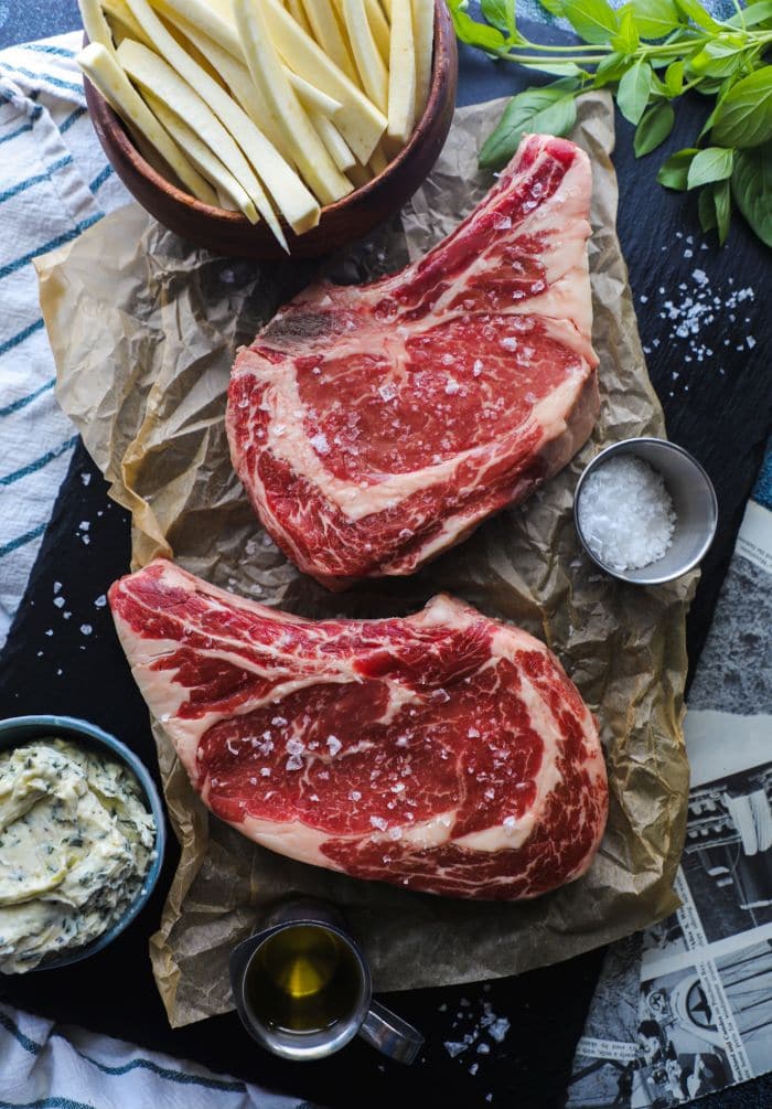 Two raw bone in ribeyes with course sea salt on a black tray. 