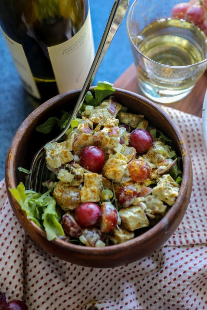 A close up of a serving of grilled chicken salad in a brown bowl with lettuce and a glass of wine. 