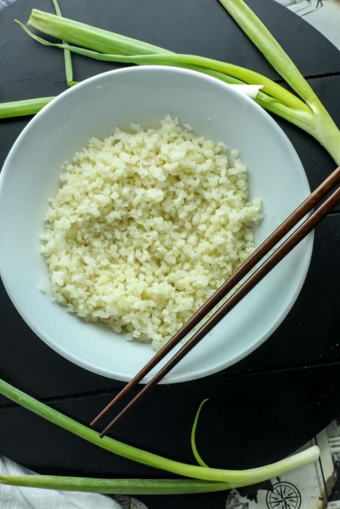 Par-cooked frozen cauliflower rice in a white bowl with brown chopsticks. 