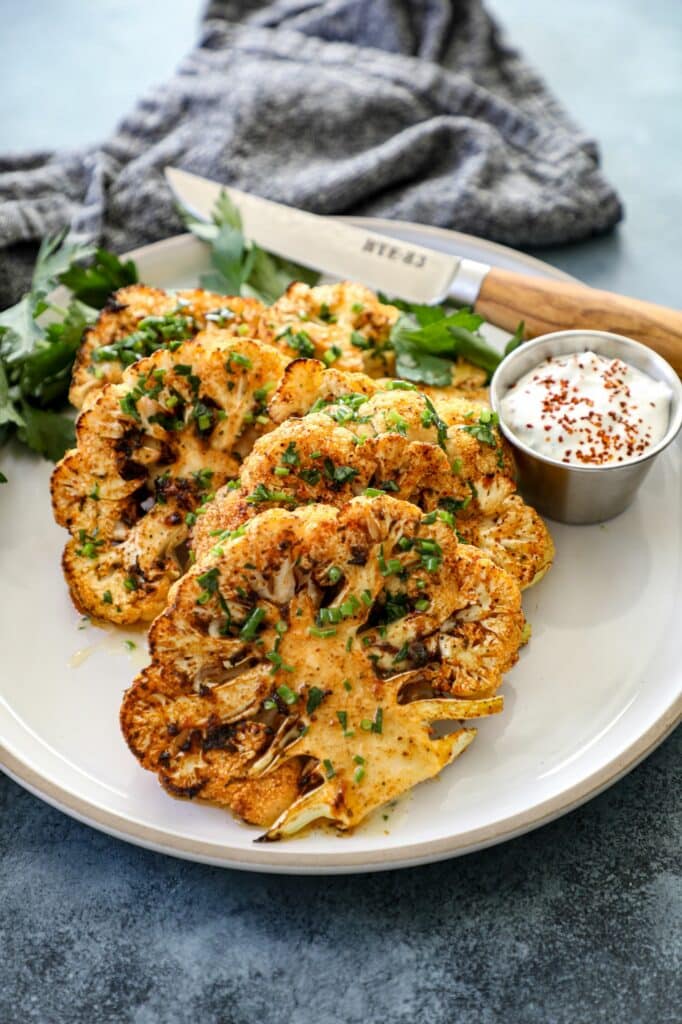 Cauliflower steaks lined up on a white plate next to sauce and a knife.