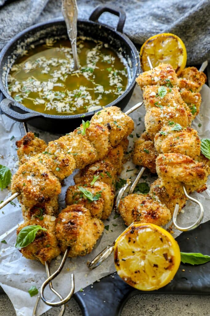 Breaded chicken skewers stacked on top of each other next to a cast iron pan with sauce in it. 