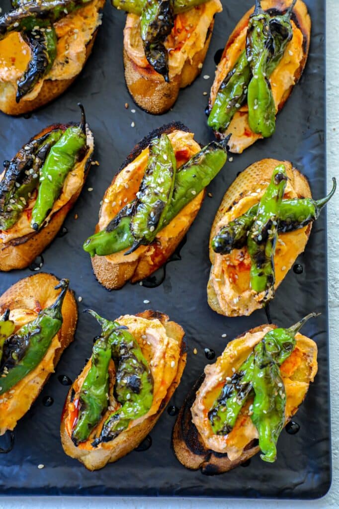 Crostini topped with grilled shishito peppers on a black serving tray. 