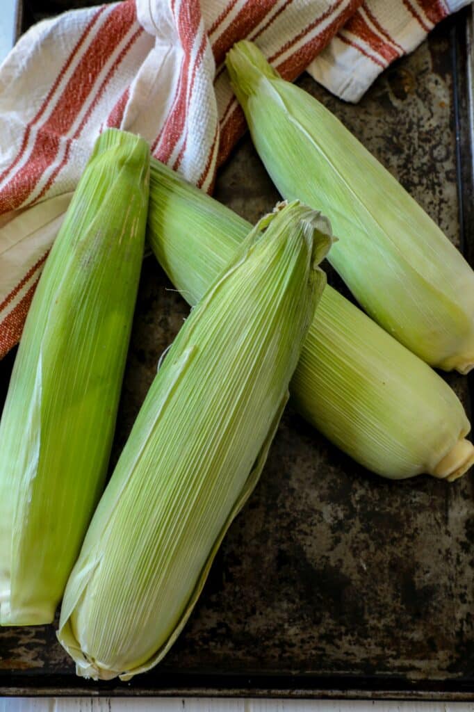 Uncooked ears of corn on a baking sheet.