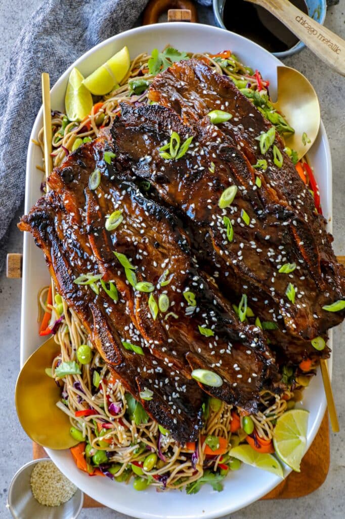 Ribs on a bed of noodles and vegetables on a white serving platter with golden serving spoons. 